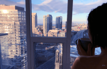 Woman drinking tea and looking at Toronto skyline from condo window.